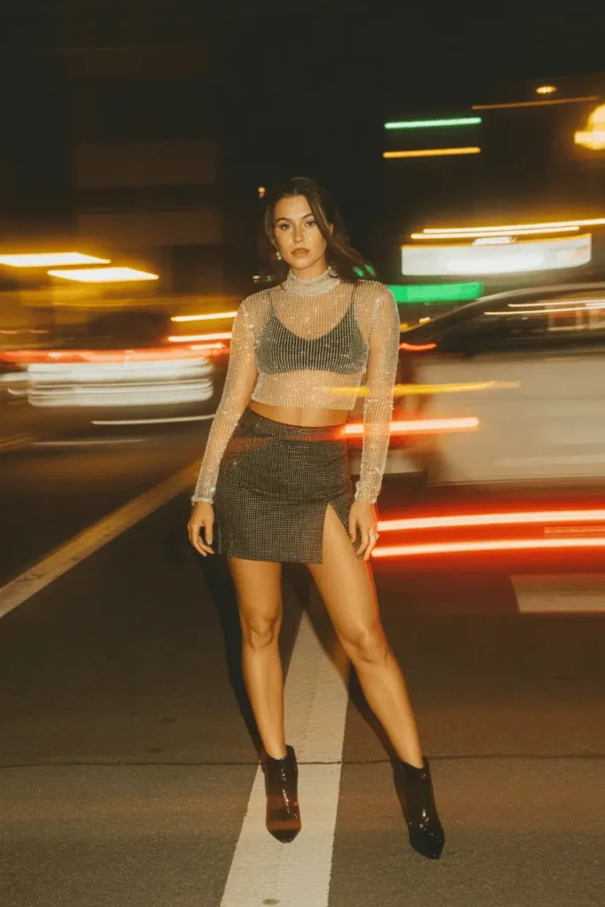 Sparkly rhinestone or sequin top paired with a mini skirt and heeled boots. Nighttime photo with blurred city lights, glamorous glow, confident pose.