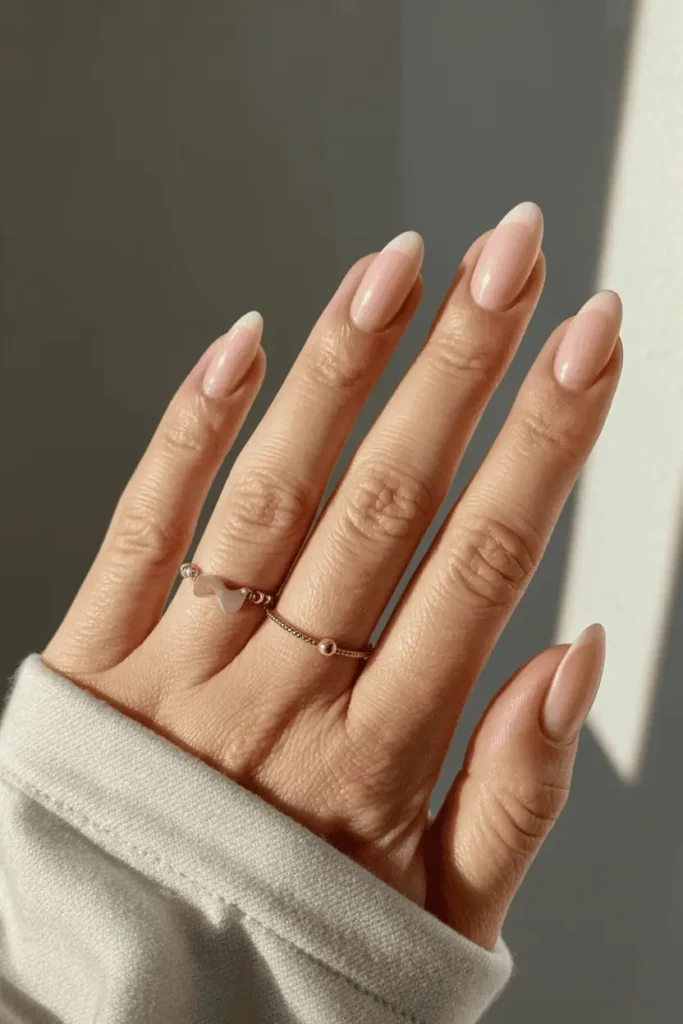 Short almond nails in soft blush pink polish. Clean daylight setting, minimalist jewelry accent.