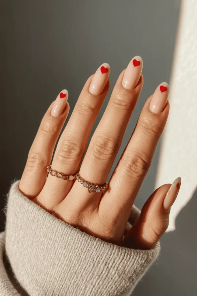 Short nude nails with small red hearts on ring fingers. Romantic cozy setup, soft warm lighting.