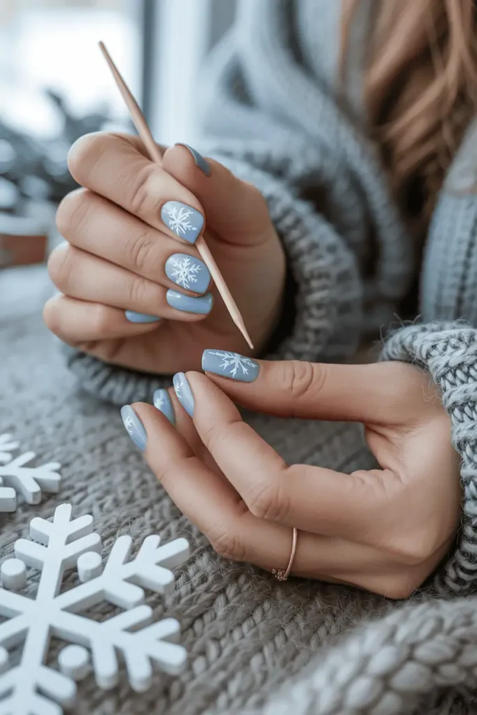 White snowflake nail art on pale blue nails, hands painting details with a toothpick, cozy winter background.
Pinterest hero photo, soft natural daylight, realistic hands, cool neutral palette, props: snowflakes, knit blanket.