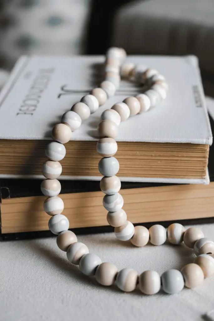 A wooden bead garland with beads painted in soft pastel spring colors—half-dipped in paint creating two-toned effects—draped across a stack of books on a coffee table. The beads show handmade paint variations. Natural light, textural styling, calm and modern aesthetic.