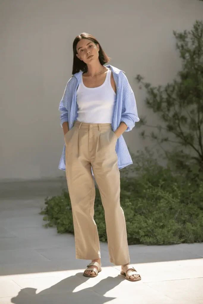 Relaxed woman wearing beige linen trousers, white tank top, and an oversized light blue button-up shirt worn open. Minimal sandals, soft sunlight, calm minimalist background with greenery.