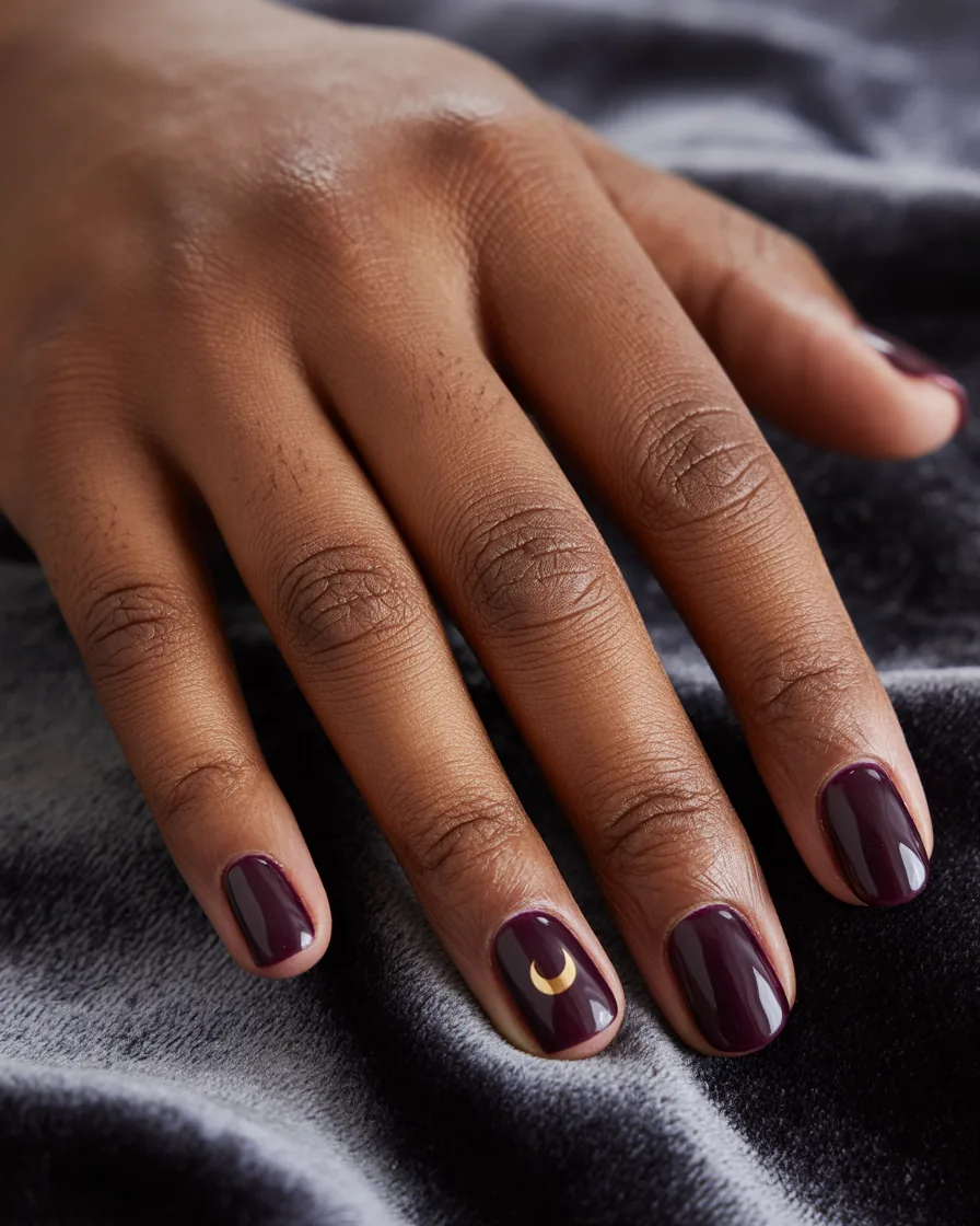 Short oval nails in glossy deep plum with a small gold crescent moon on the ring finger
